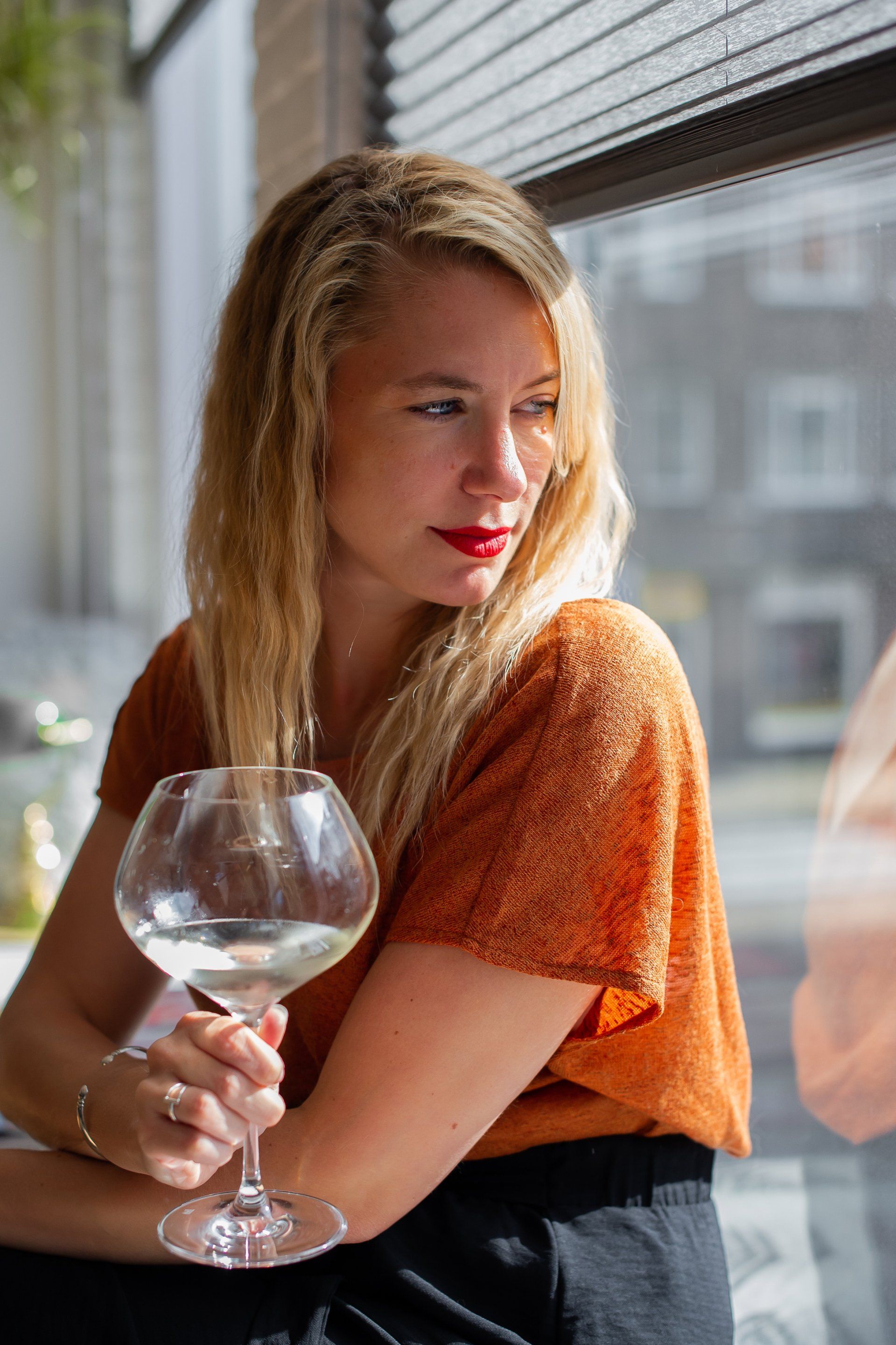 Vrouw met blond haar, die een wijnglas vasthoudt en uit het raam kijkt. Ze draagt ​​een oranje shirt en rode lippenstift.