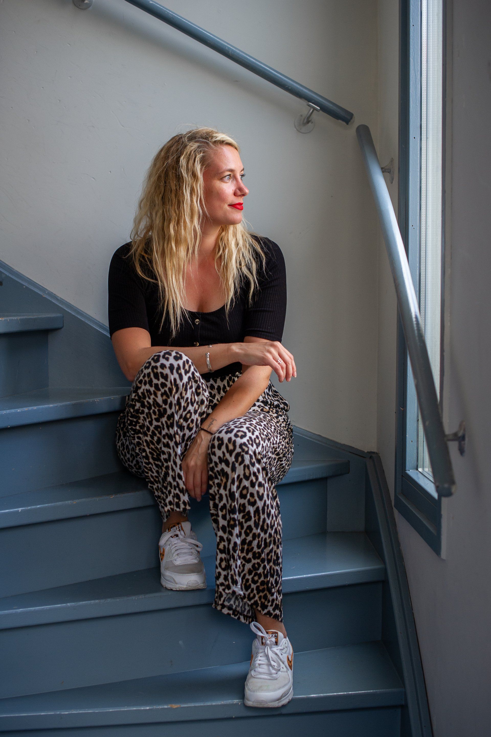 Een vrouw met krullend blond haar zit op een blauwe trap en kijkt uit het raam. Ze draagt ​​een zwart shirt, een broek met luipaardprint en witte sneakers.