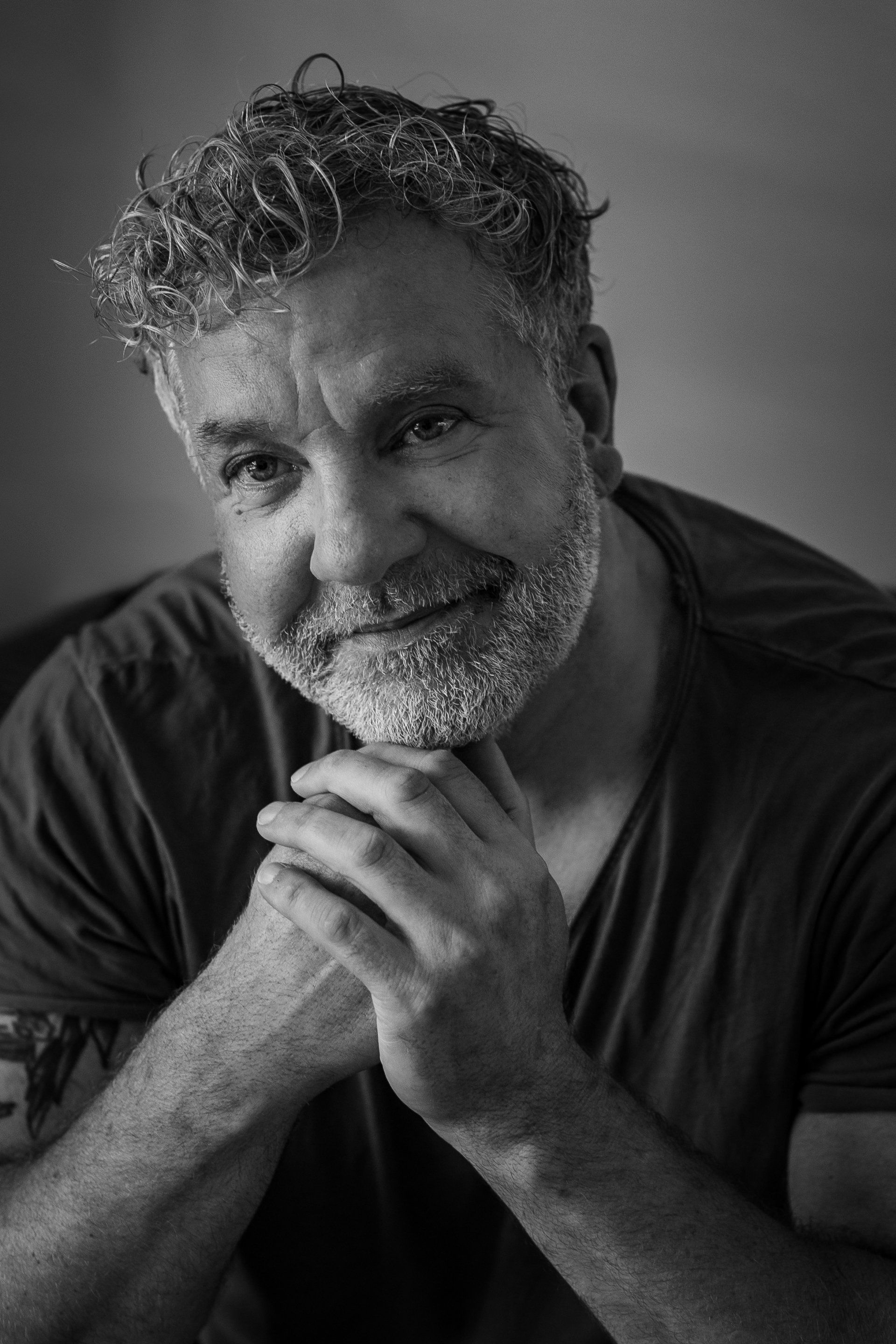 Man met grijs wordend haar en baard, handen ineengevouwen, licht glimlachend.