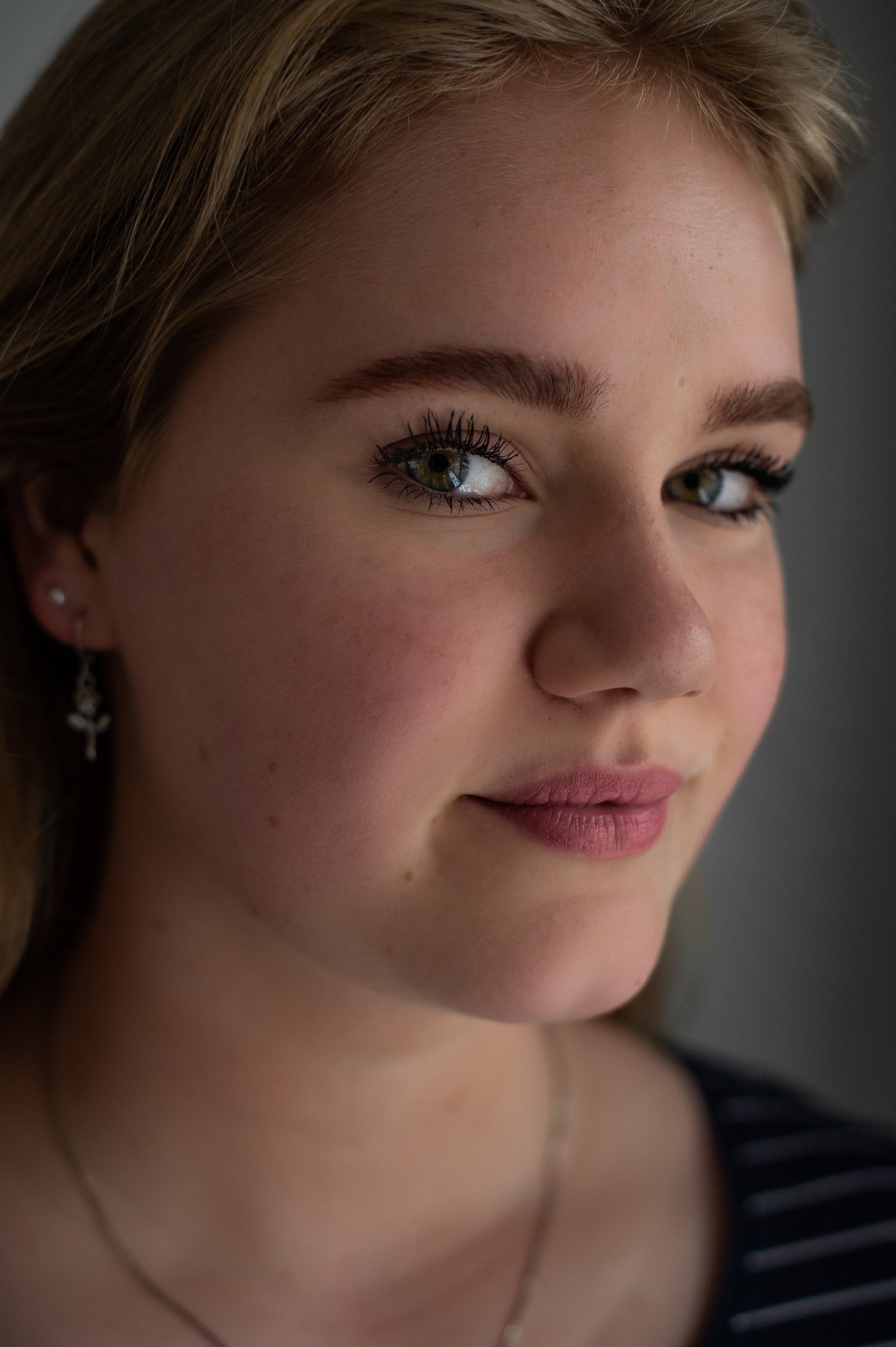 Blondine met blauwe ogen en een lichte blos kijkt naar de camera, draagt ​​kleine oorbellen, lacht en heeft donkere mascara op.