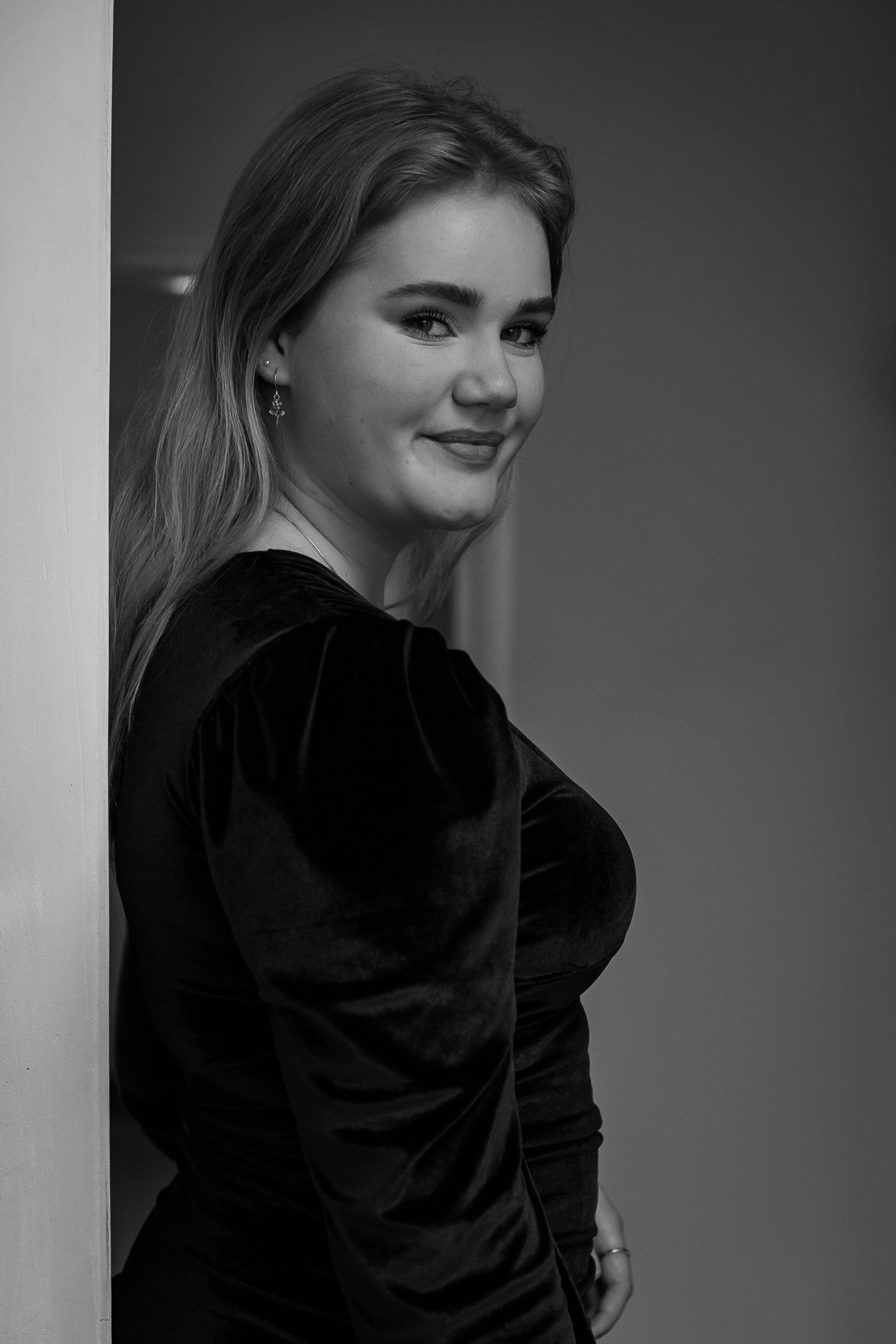 Vrouw in een zwarte jurk leunt lachend tegen een muur. Zwart-witfoto.