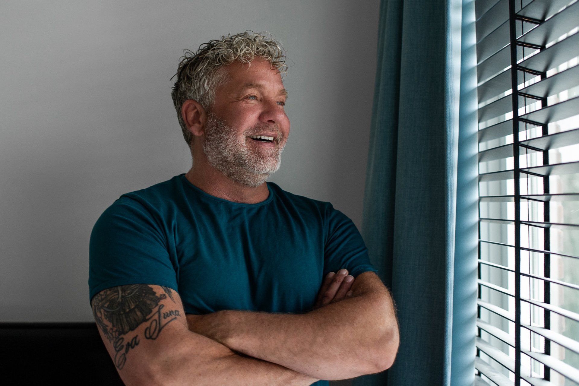 Een man met grijs krullend haar glimlacht terwijl hij uit het raam kijkt, waar de jaloezieën gesloten zijn. Hij heeft zijn armen over elkaar en draagt ​​een blauw shirt.