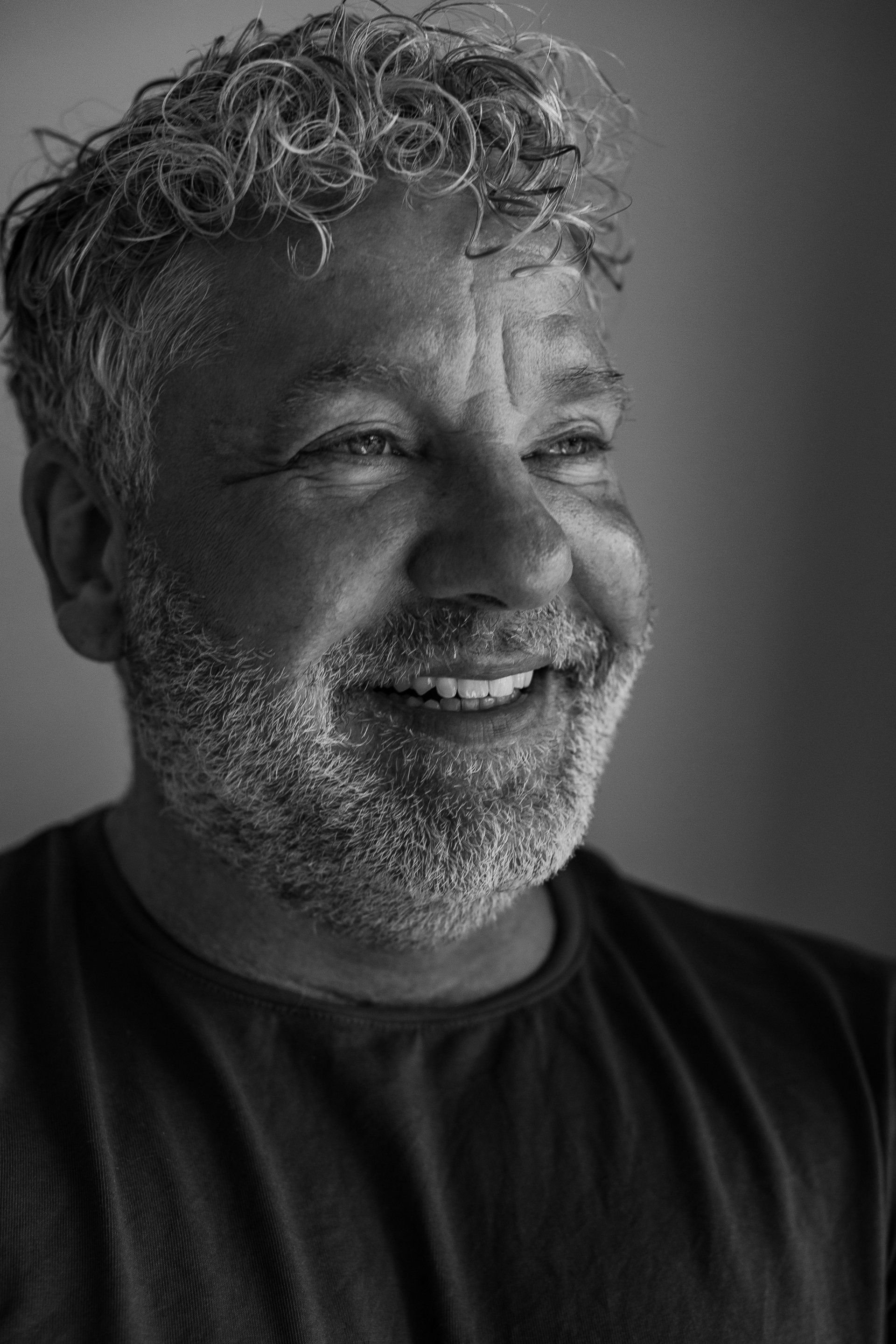 Man met krullend haar en baard lacht breeduit, portret van hoofd en schouders, zwart-wit.