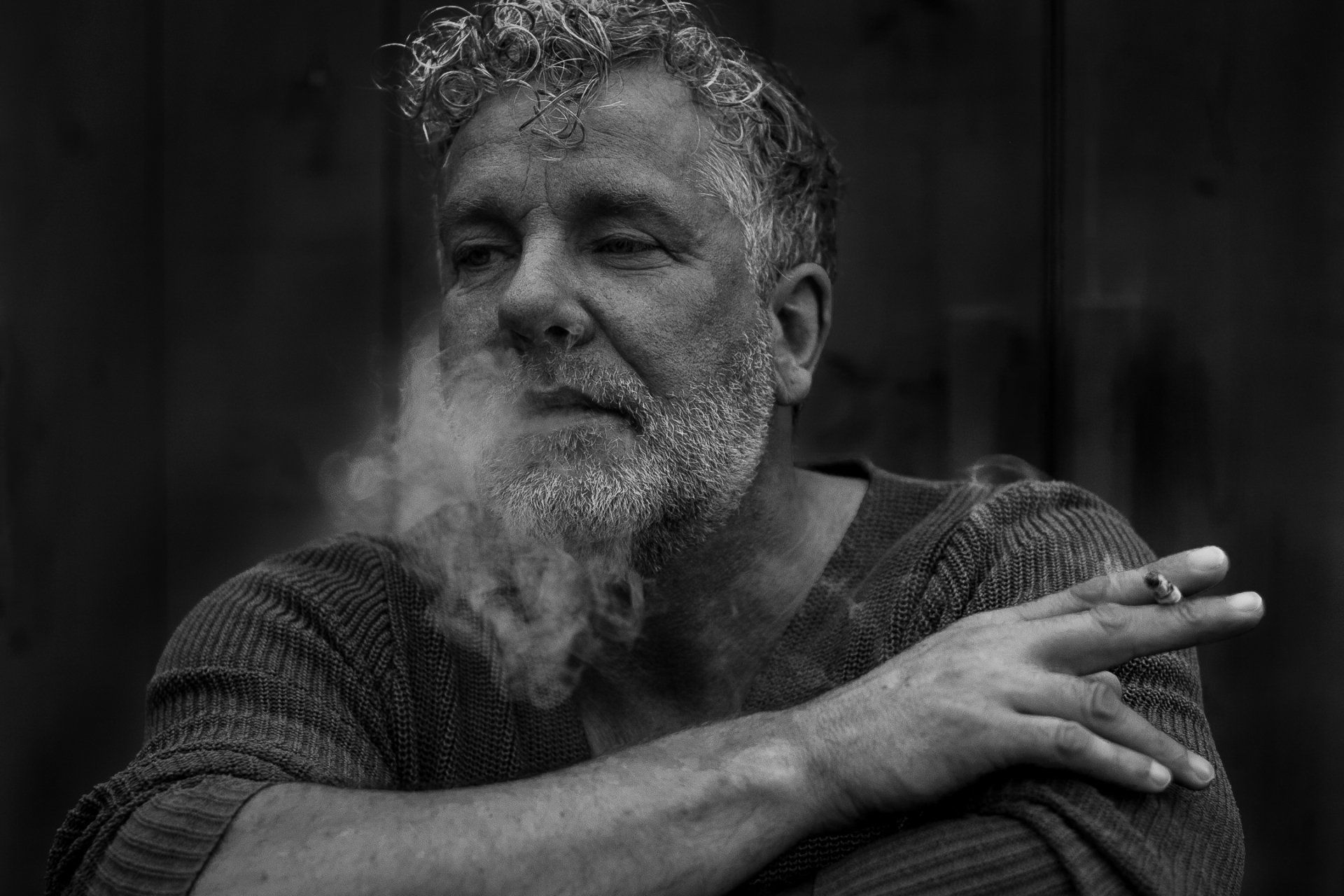 Een man met een baard blaast sigarettenrook uit, zittend in een zwart-witfoto in de buitenlucht.