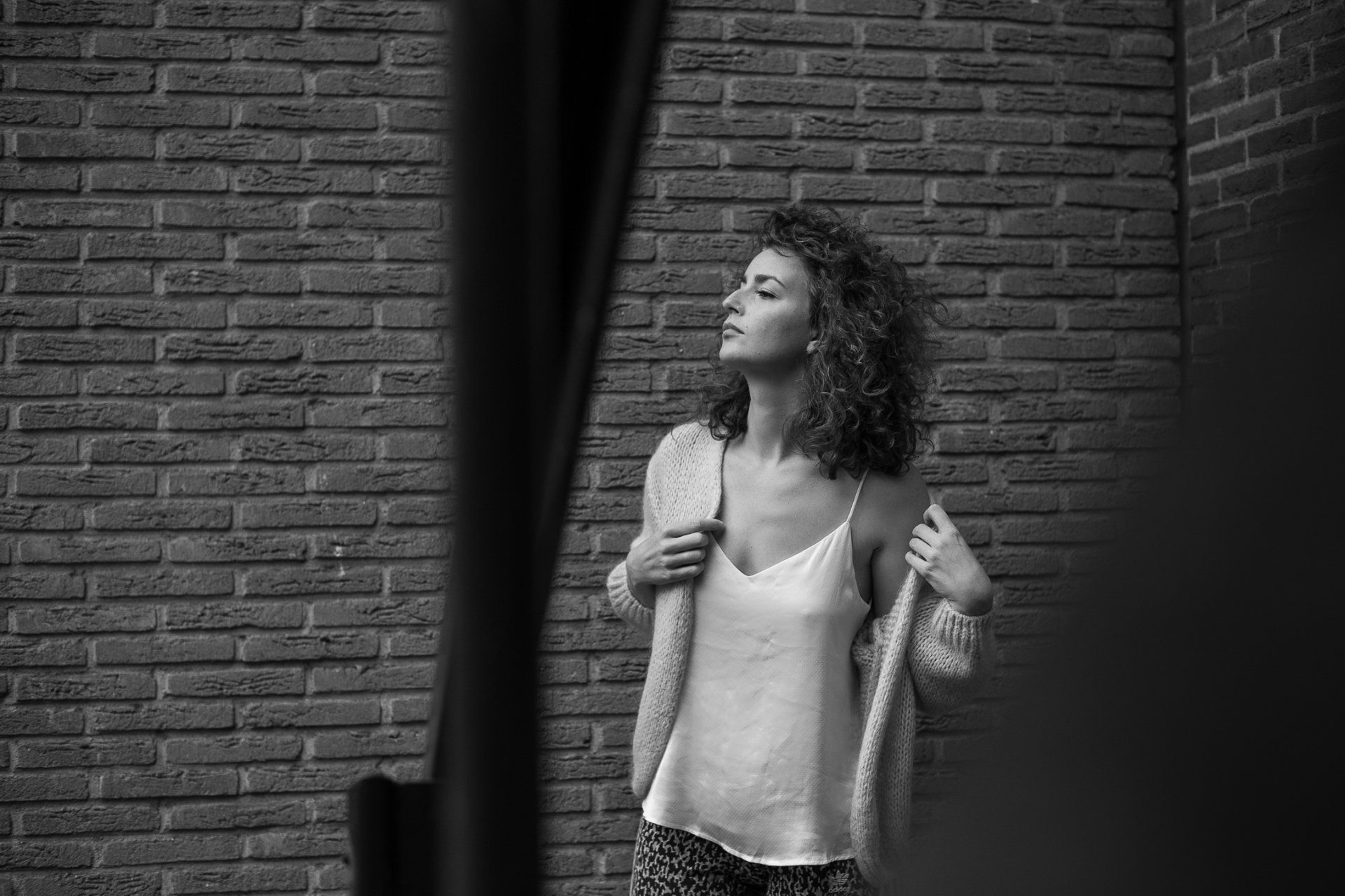Vrouw in een hemdje en trui, leunend tegen een bakstenen muur, kijkt weg.
