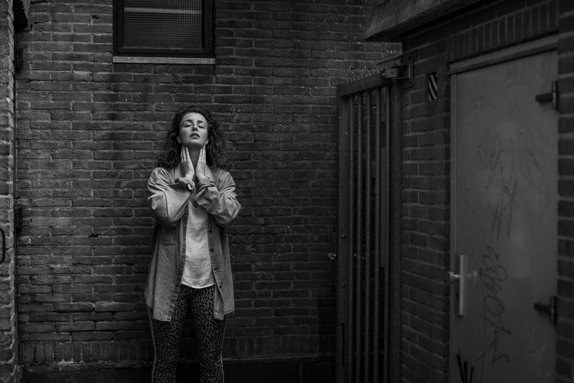 Een vrouw in een broek en jas met patroon staat tegen een bakstenen muur in een steegje, met haar handen ineengevouwen bij haar gezicht.