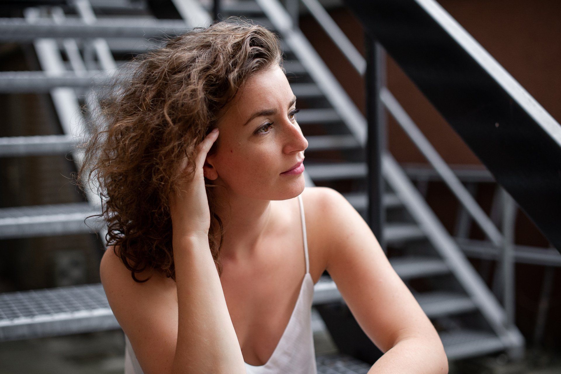 Een vrouw met krullend haar zit op een metalen trap en kijkt niet in beeld, terwijl ze met haar hand haar haar aanraakt.