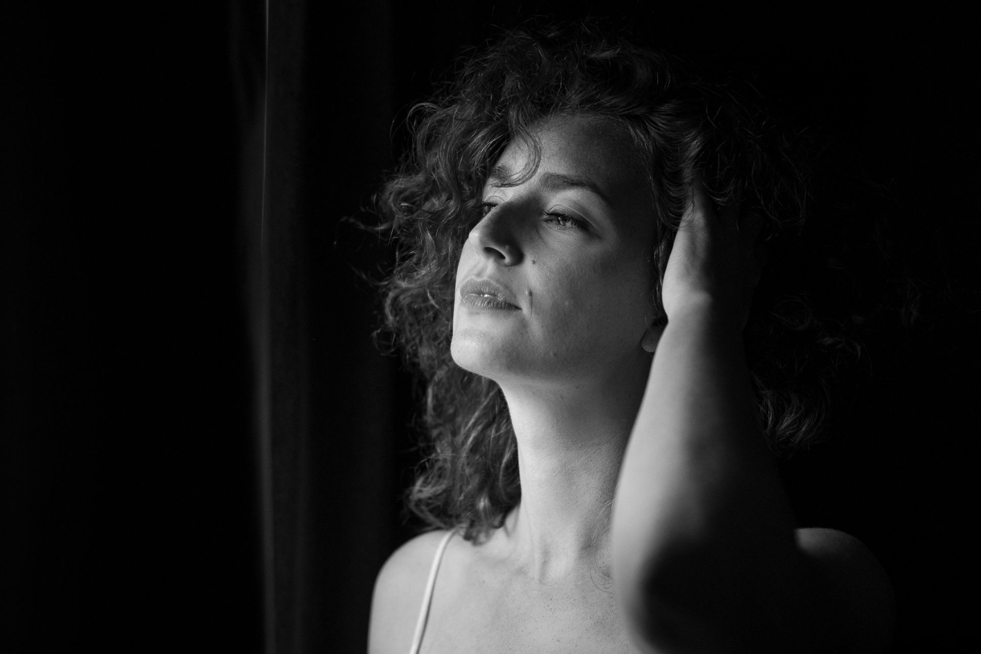 Vrouw met krullend haar, in de schaduw, kijkt omhoog, hand in haar haar.