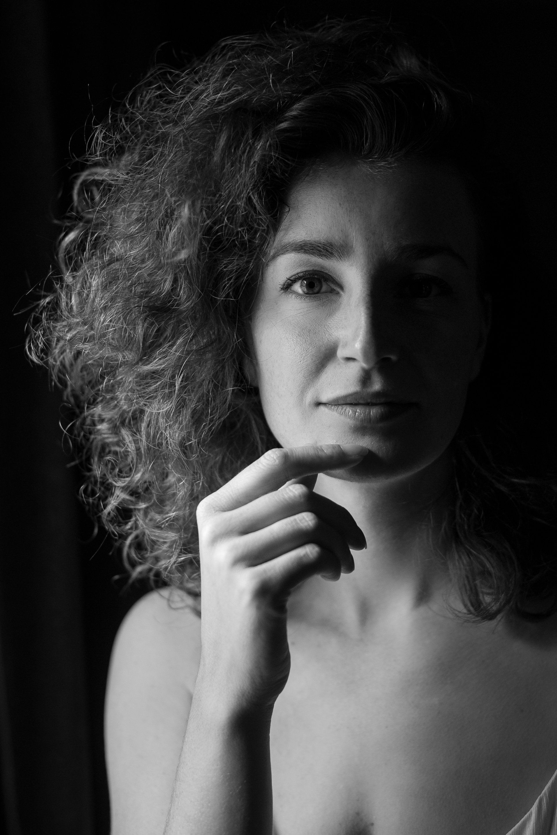 Vrouw met krullend haar, hand aan haar kin, in een donkere omgeving, kijkt naar de kijker.