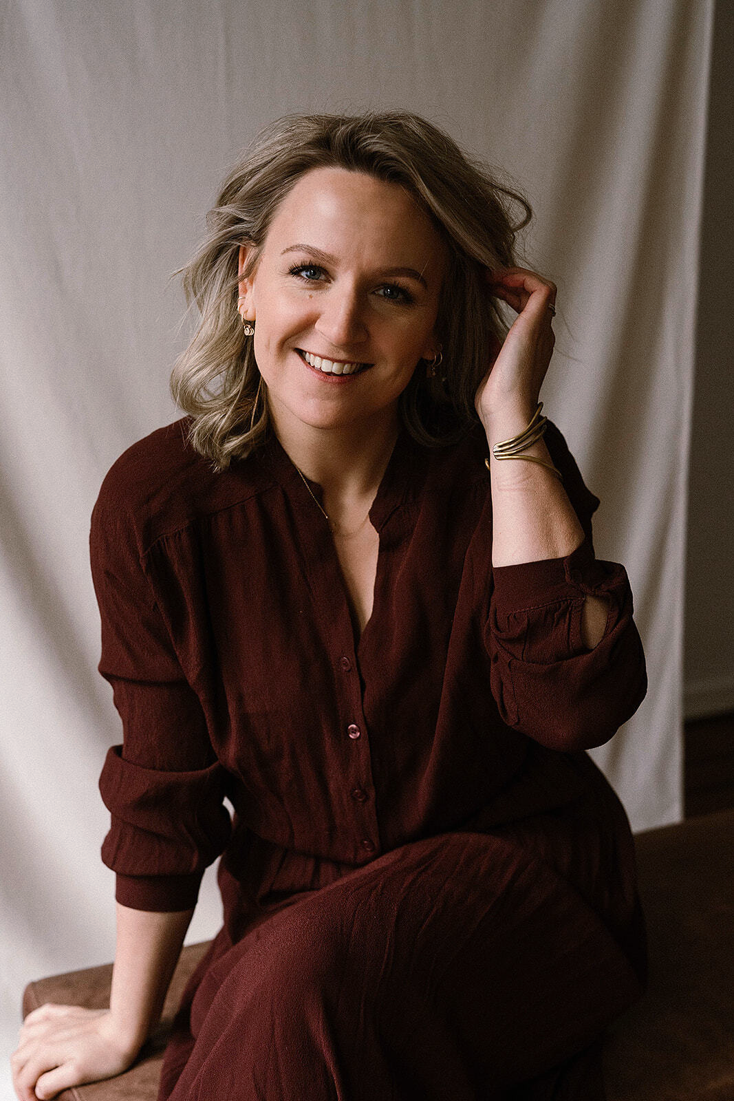 Een vrouw met krullend haar, gekleed in een bordeauxrode blouse en broek, lacht en heeft haar hand in haar haar.