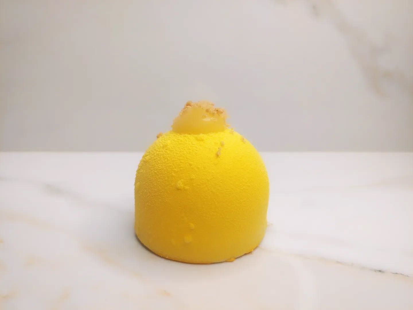 Dolce giallo a forma di cupola, possibilmente una mousse, con glassa e una piccola guarnizione, su una superficie bianca.