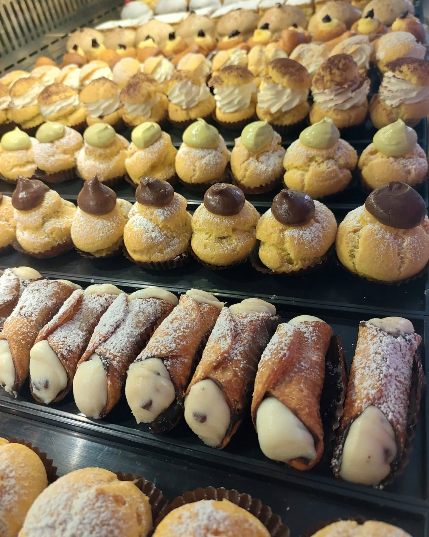 Pasticcini esposti in vetrina: bignè, cannoli e altri piccoli dessert, alcuni spolverati con zucchero a velo.