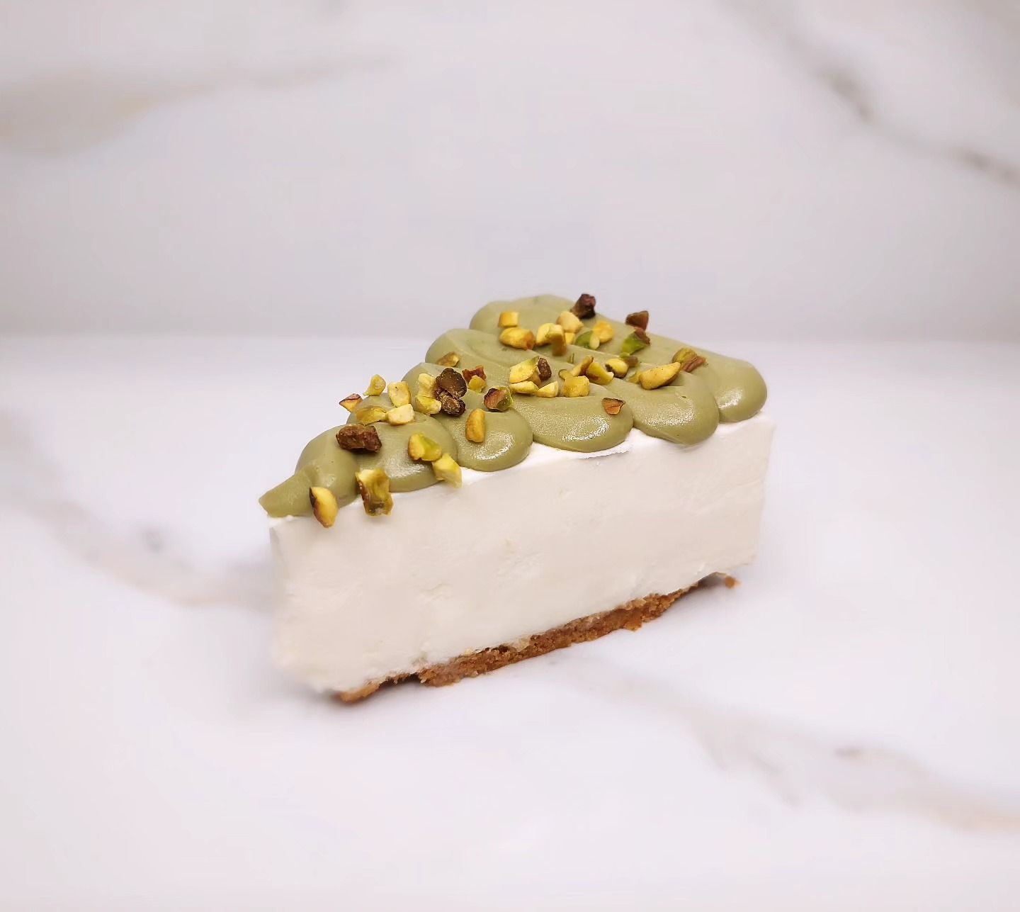 Fetta di cheesecake con copertura al pistacchio su una superficie bianca.