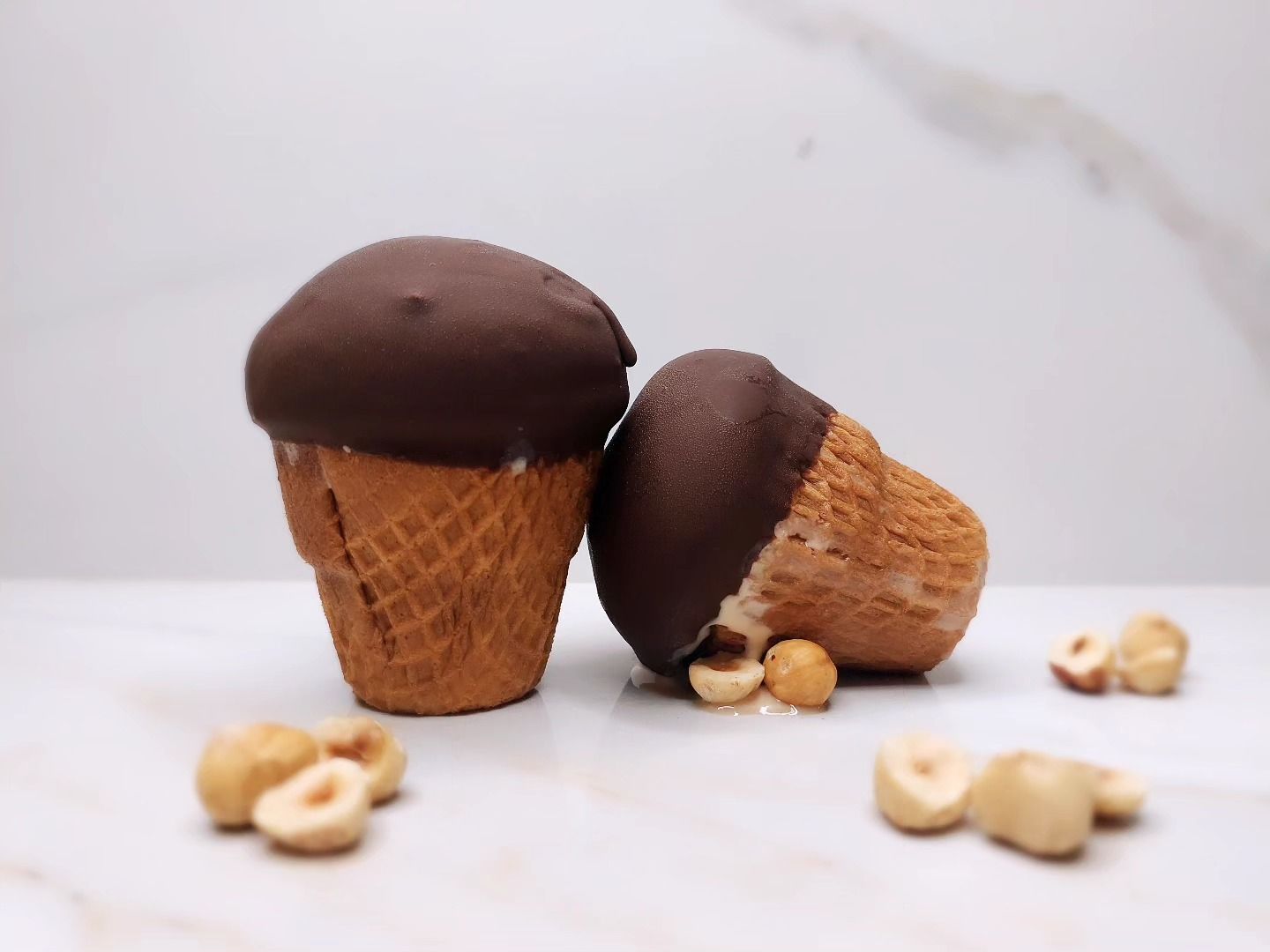 Due coni gelato ricoperti di cioccolato con nocciole sparse su una superficie di marmo.