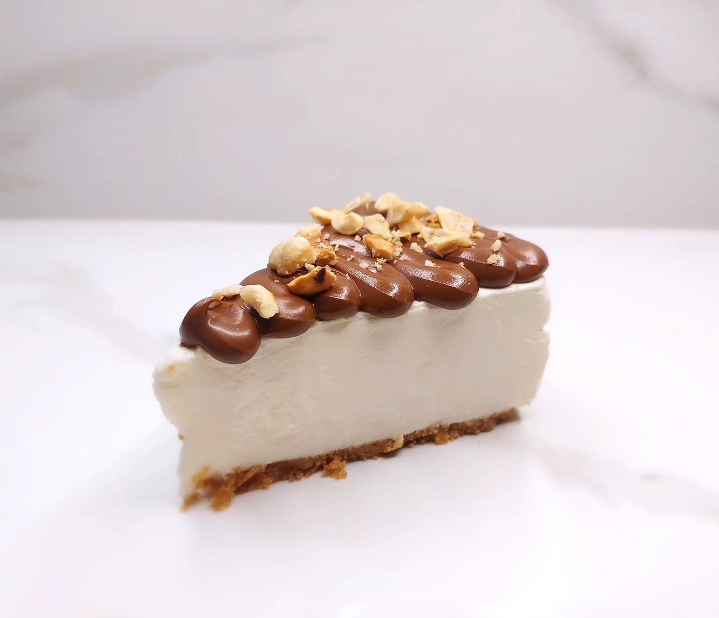 Fetta di cheesecake con copertura al cioccolato e noci tritate su una superficie bianca.