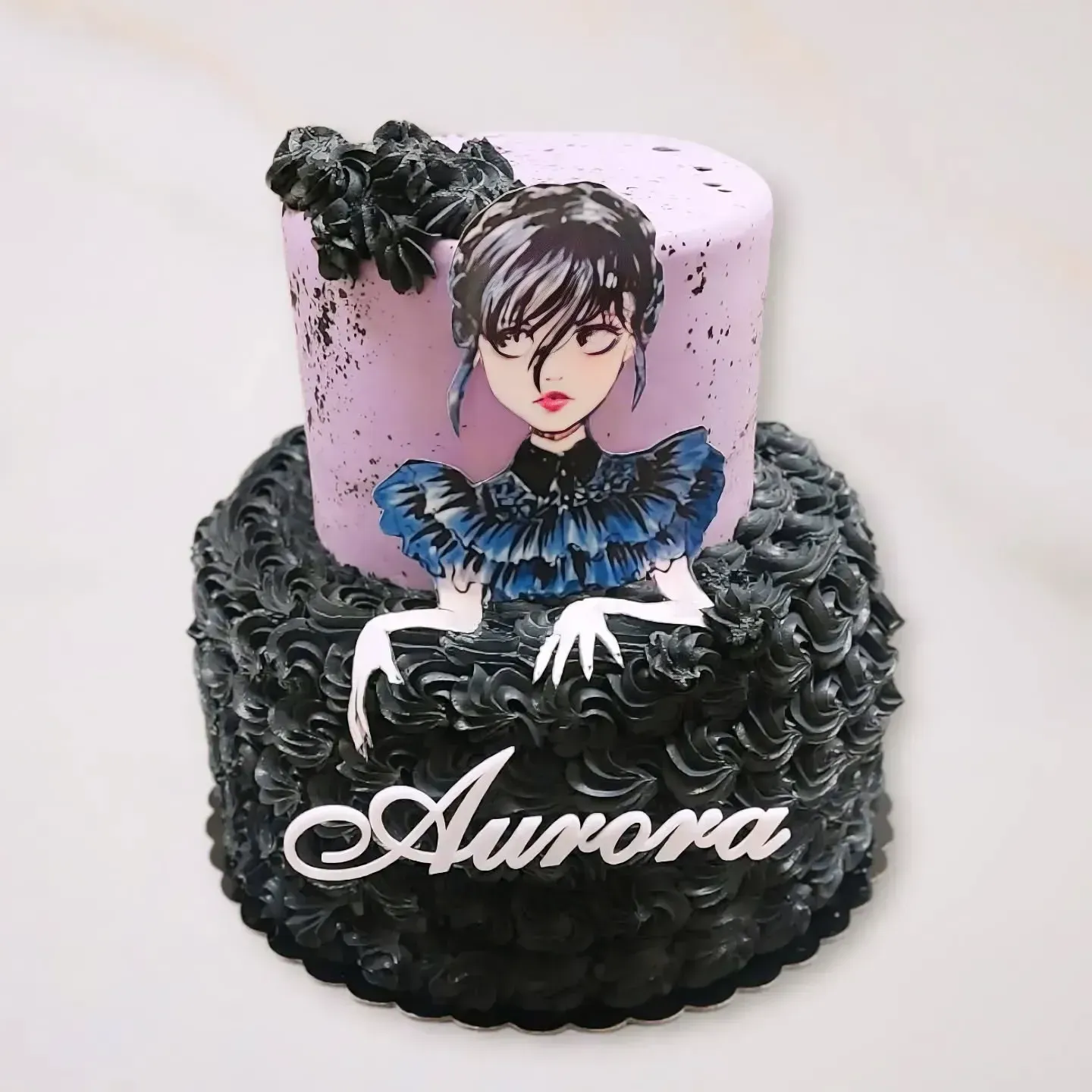 Torta a due piani con glassa nera e piano superiore lilla con un disegno di Mercoledì Addams e la scritta 