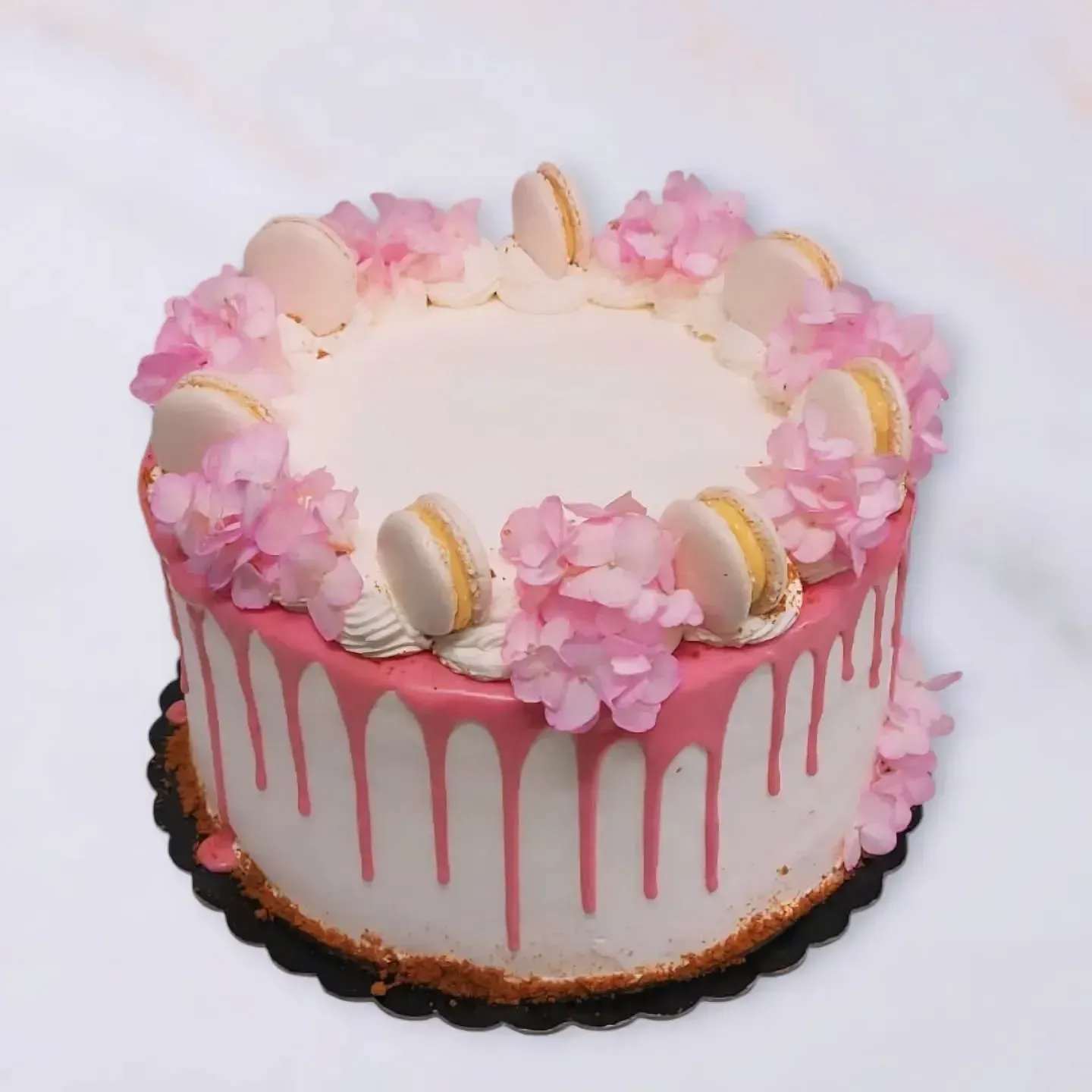 Torta con glassa rosa, decorata con fiori rosa e macaron, su un'alzata nera.