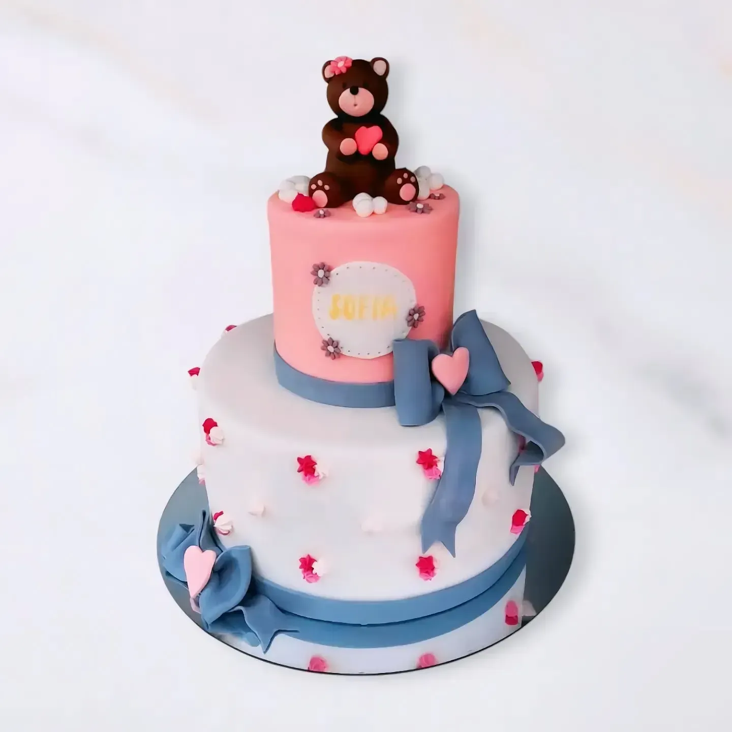 Torta a due piani con glassa rosa e bianca, fiocco, decorazioni floreali e un orsacchiotto in cima.