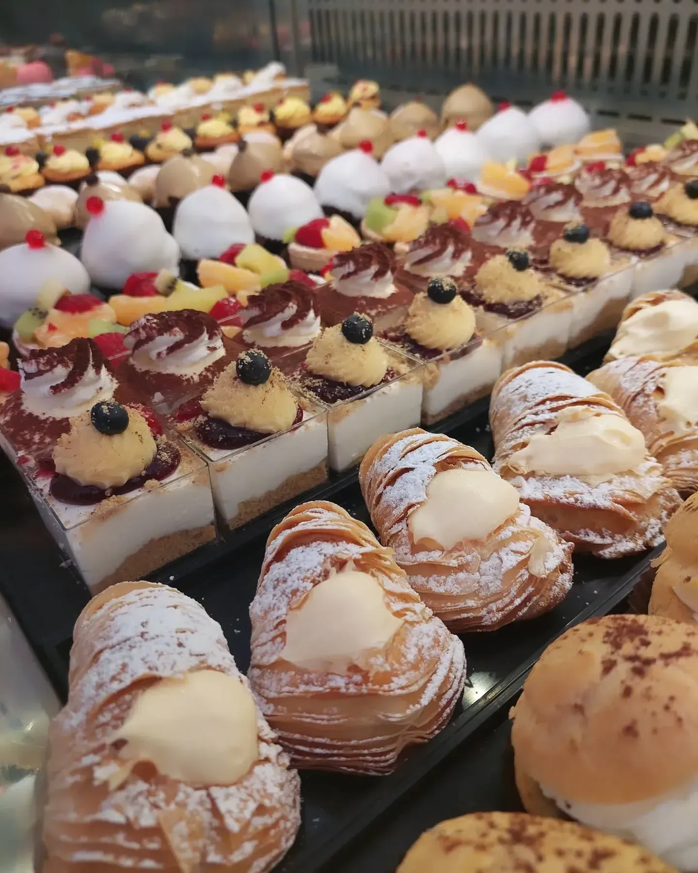 Pasticcini esposti in una vetrina: torte, bignè e crostate con varie guarnizioni e zucchero a velo.