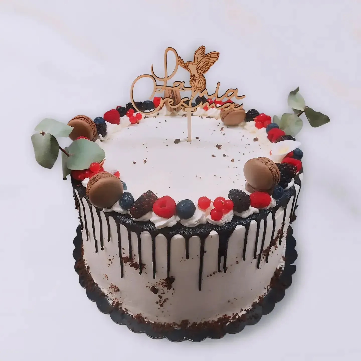 Torta decorata con frutti di bosco, gocce di cioccolato, macaron e una decorazione in legno.