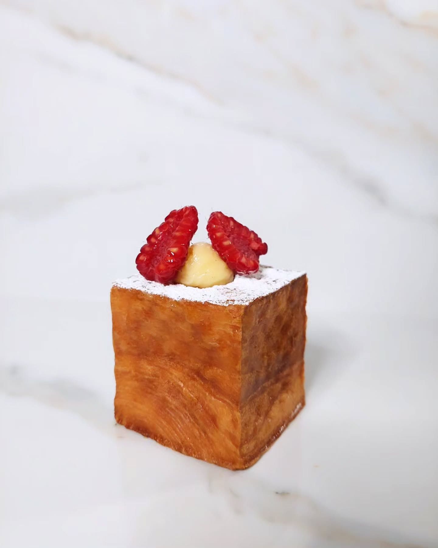 Pasta sfoglia a forma di cubo con strati e guarnizione di lamponi su una superficie di marmo bianco.