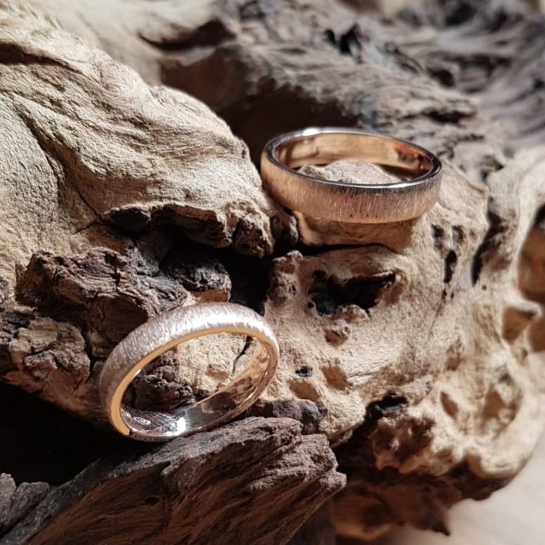 Due fedi nuziali in oro rosa testurizzato poggiano su legno galleggiante.