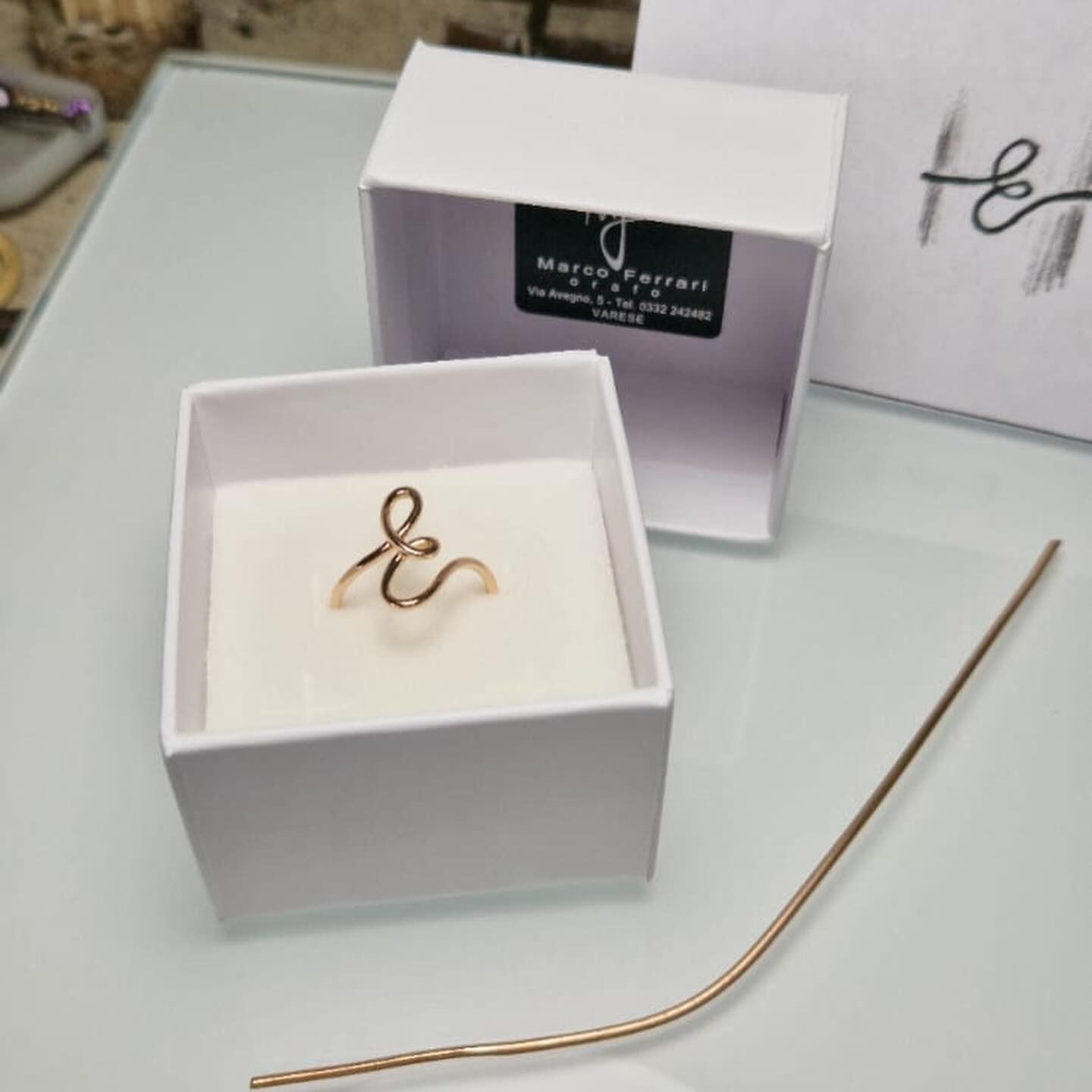 Anello d'oro a forma di e commerciale in una scatola bianca con un logo, accanto al quale è appoggiato un filo d'oro.