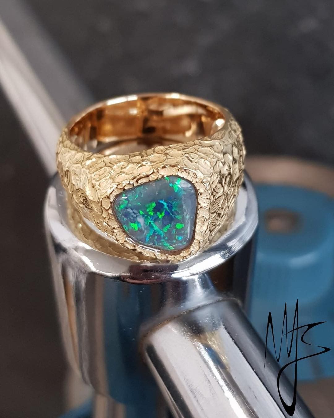 Anello in oro dalla texture ruvida, incastonato con un opale scuro che presenta riflessi verdi e blu, su una superficie metallica.