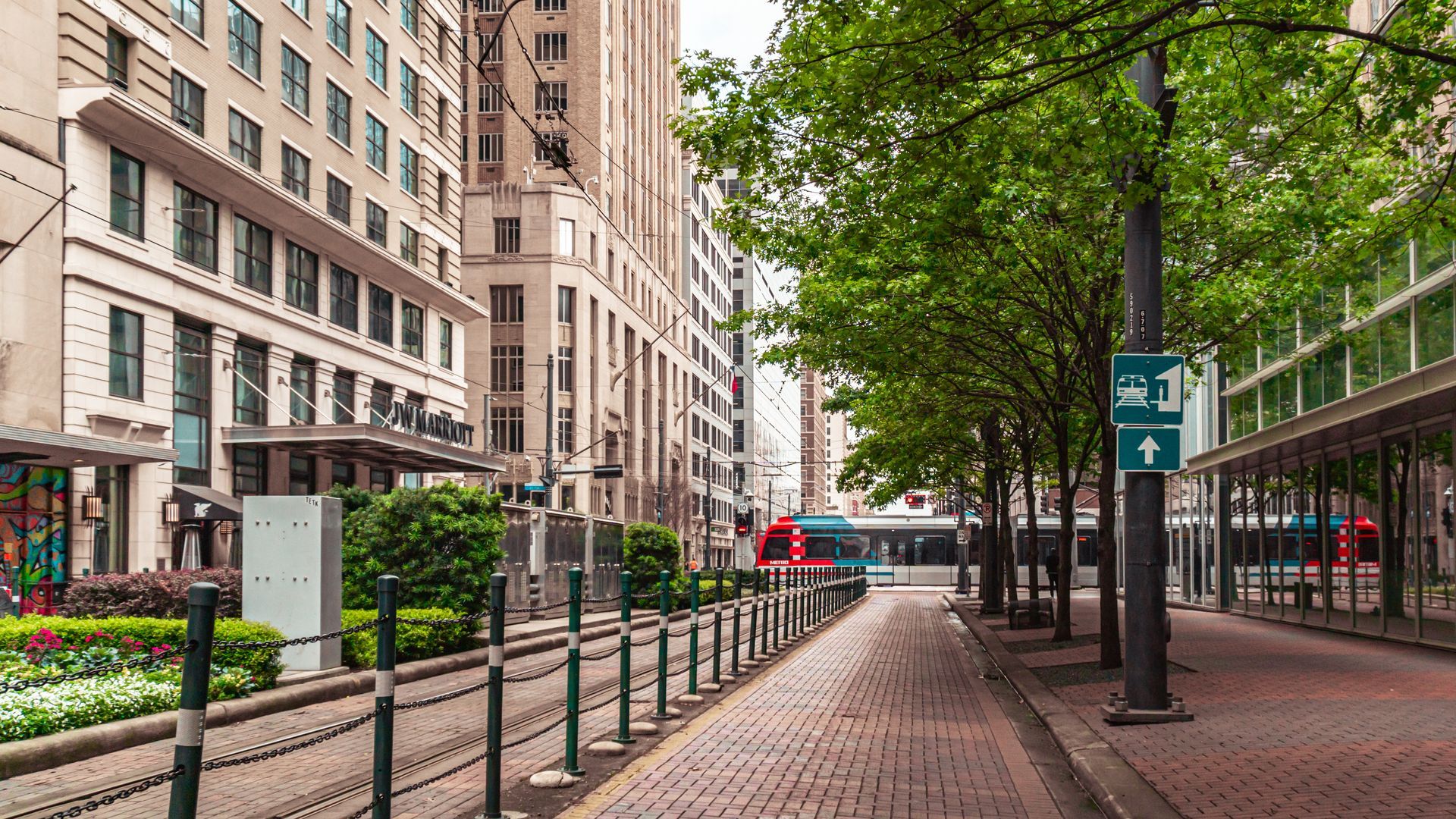 Brick-paved walkway lined with trees and buildings in a city. Tracks for a trolley run alongside the path.