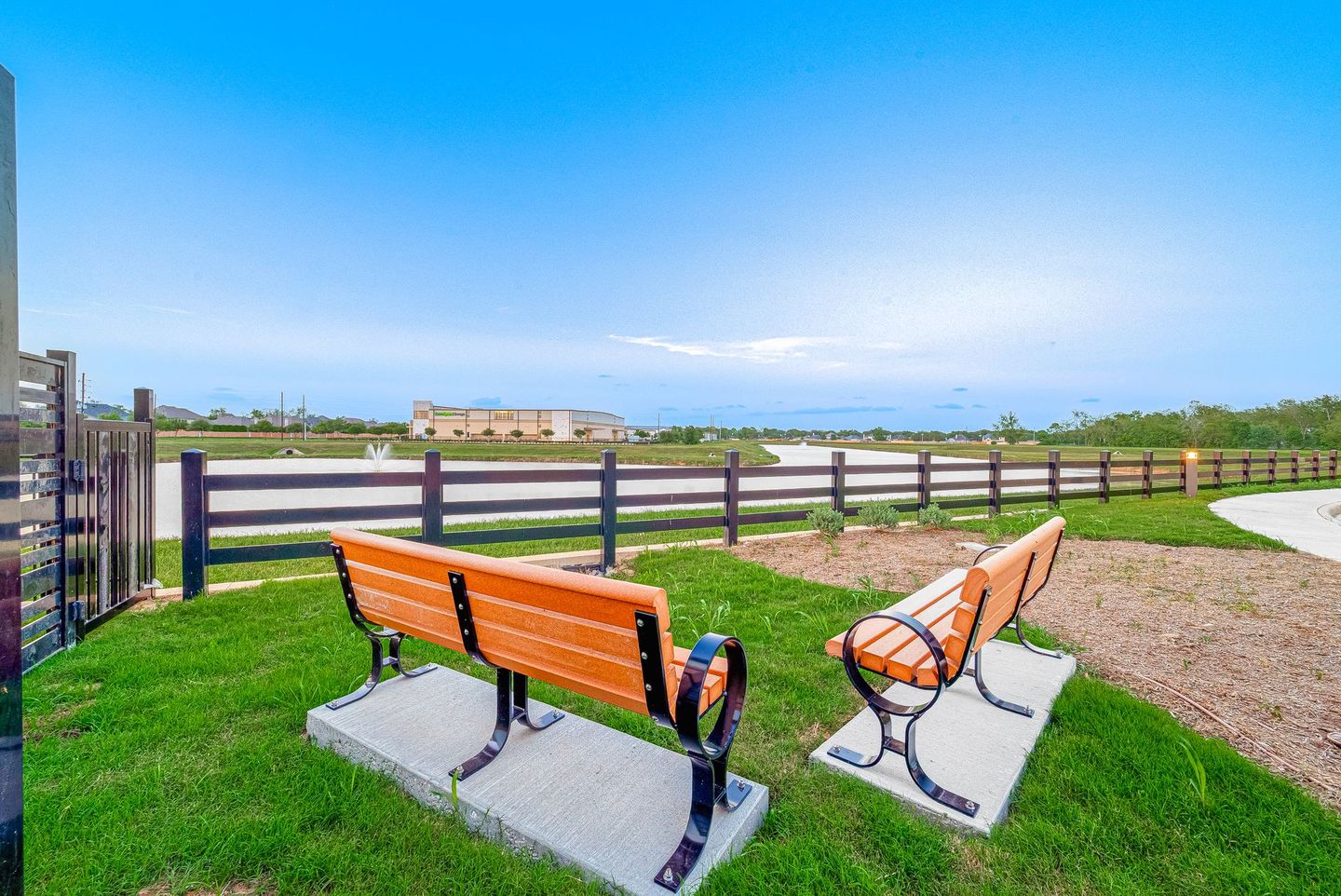 Orange benches on a grassy park overlook a fence, path, and blue water under a clear sky