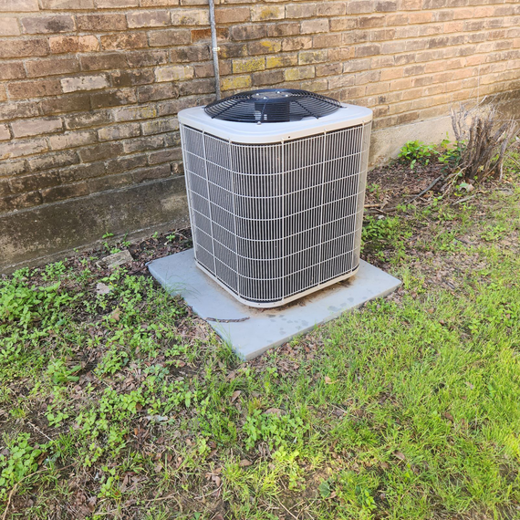 A large air conditioner is sitting in the grass next to a brick wall.