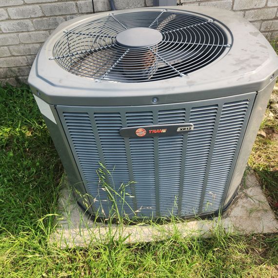 An air conditioner is sitting in the grass next to a brick wall.