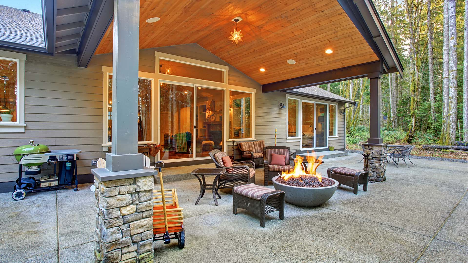 Covered patio with seating area, fire pit, and grill; house in background, wooded area.