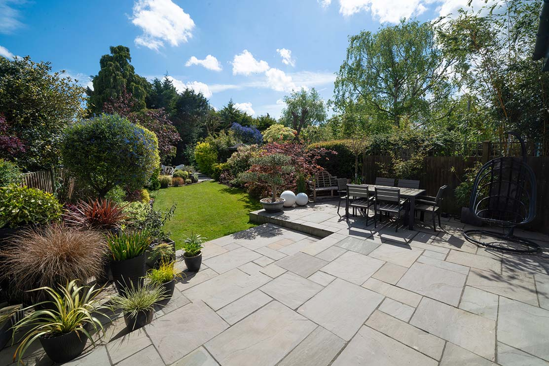 Patio with stone pavers, table, and chairs. Lush garden with green grass, various plants, and blue sky.