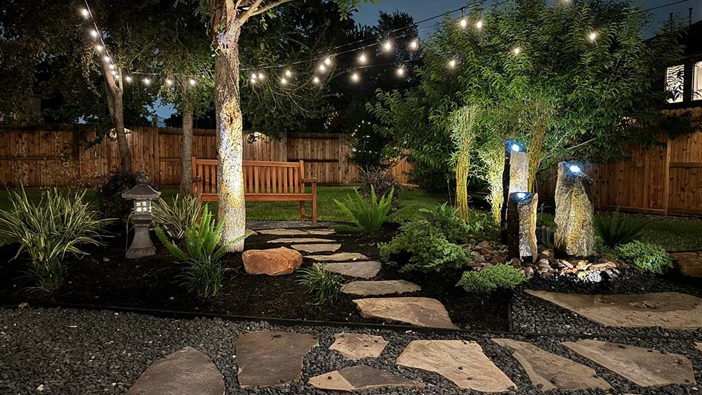 Backyard with string lights, stone path, wooden bench, and small waterfall. Nighttime setting with green plants.