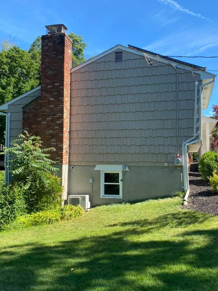 Side view of a house with grey siding, brick chimney, and a window in a grassy yard under a blue sky.