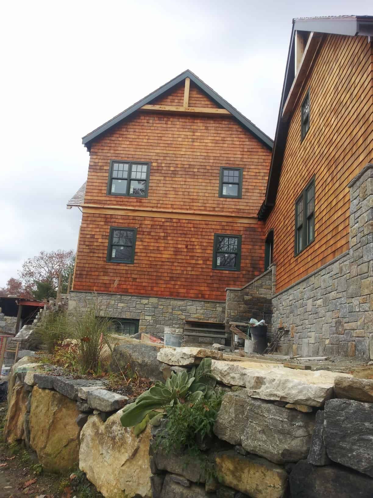 Two-story wood-shingled building with stone base, gray windows, and a stone retaining wall in front.