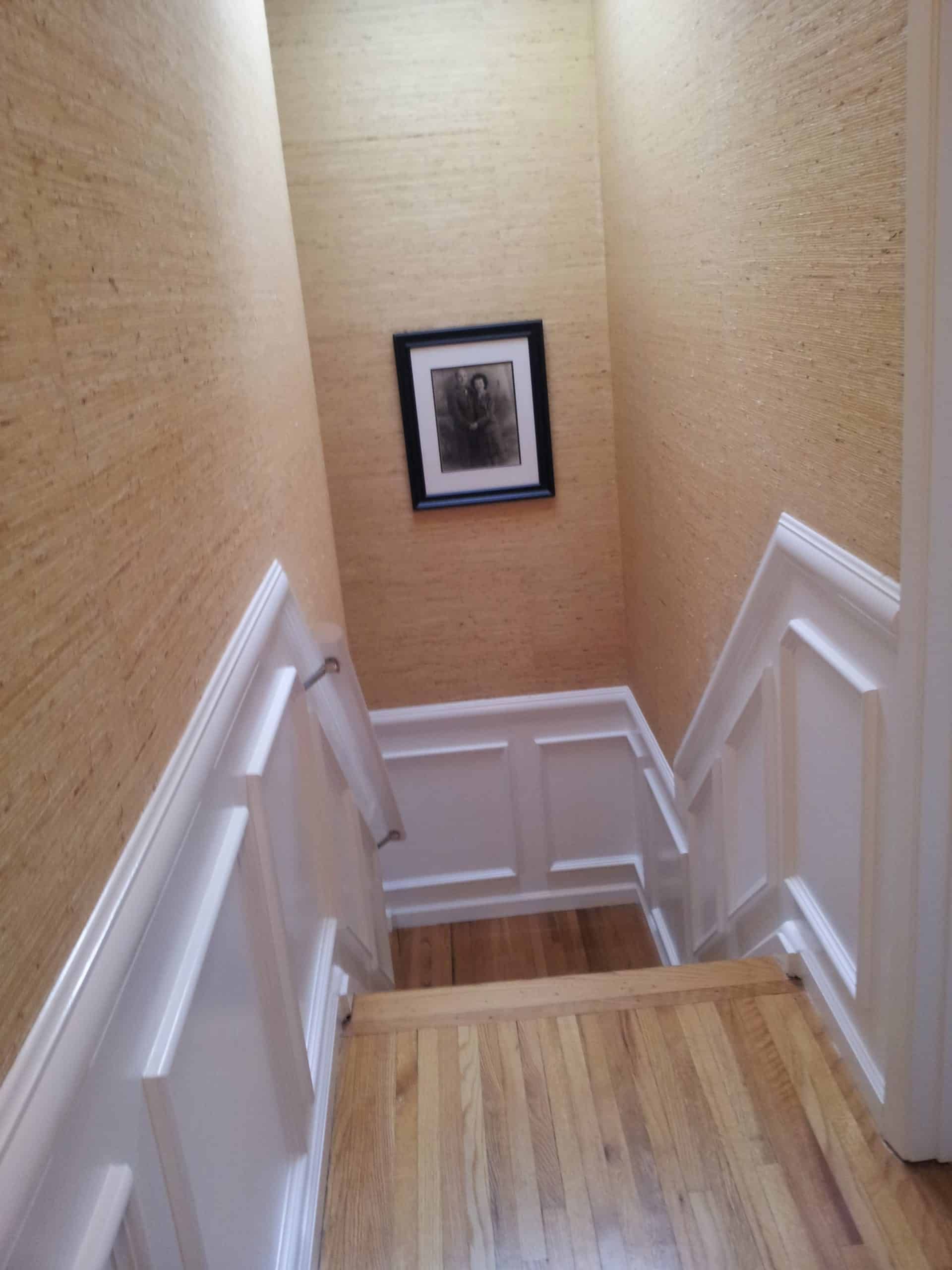 Staircase with light wood floors, white paneled walls, and beige textured wallpaper. Photo in black frame.