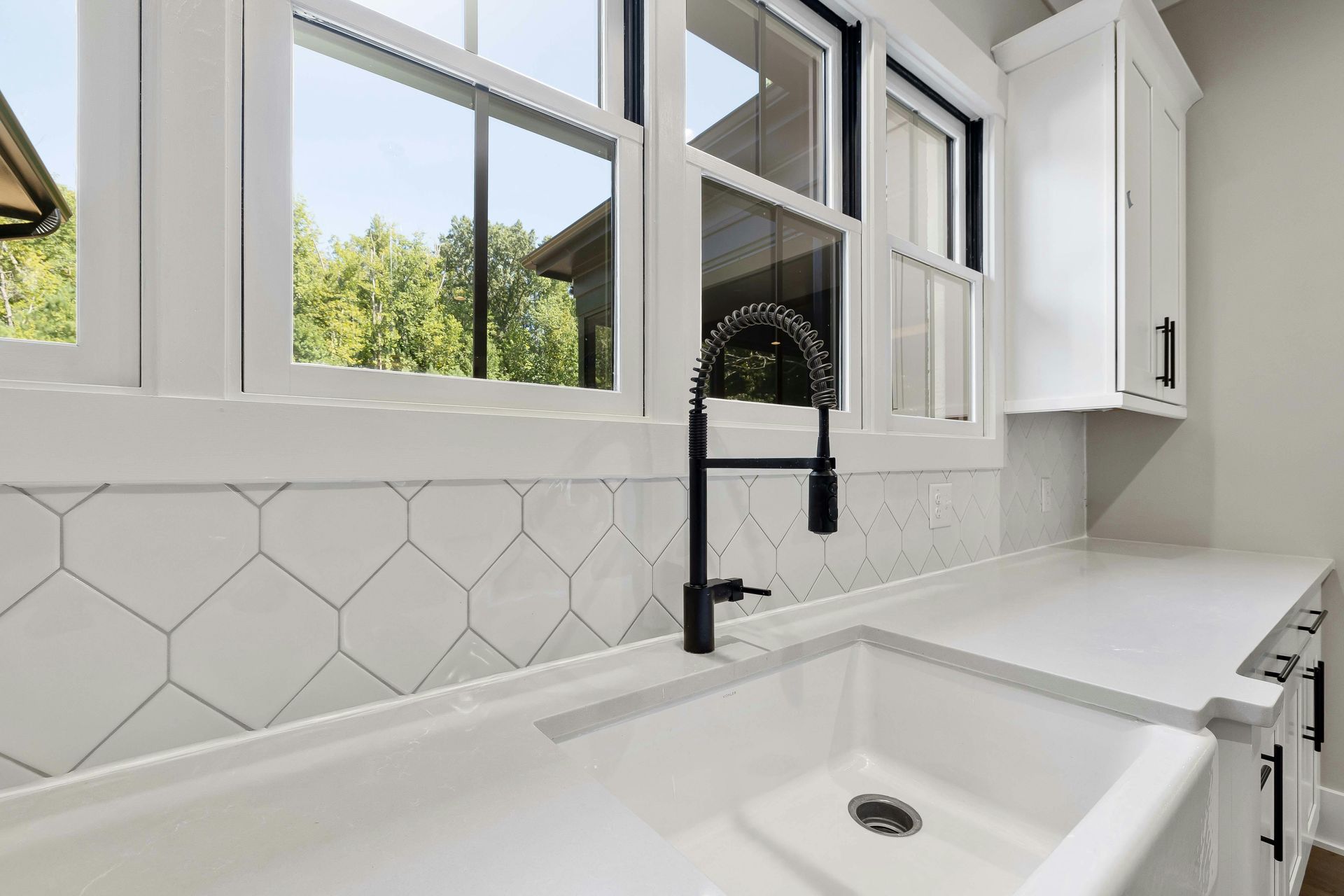 White kitchen with black faucet, geometric tile backsplash, and countertop. Window view of trees.