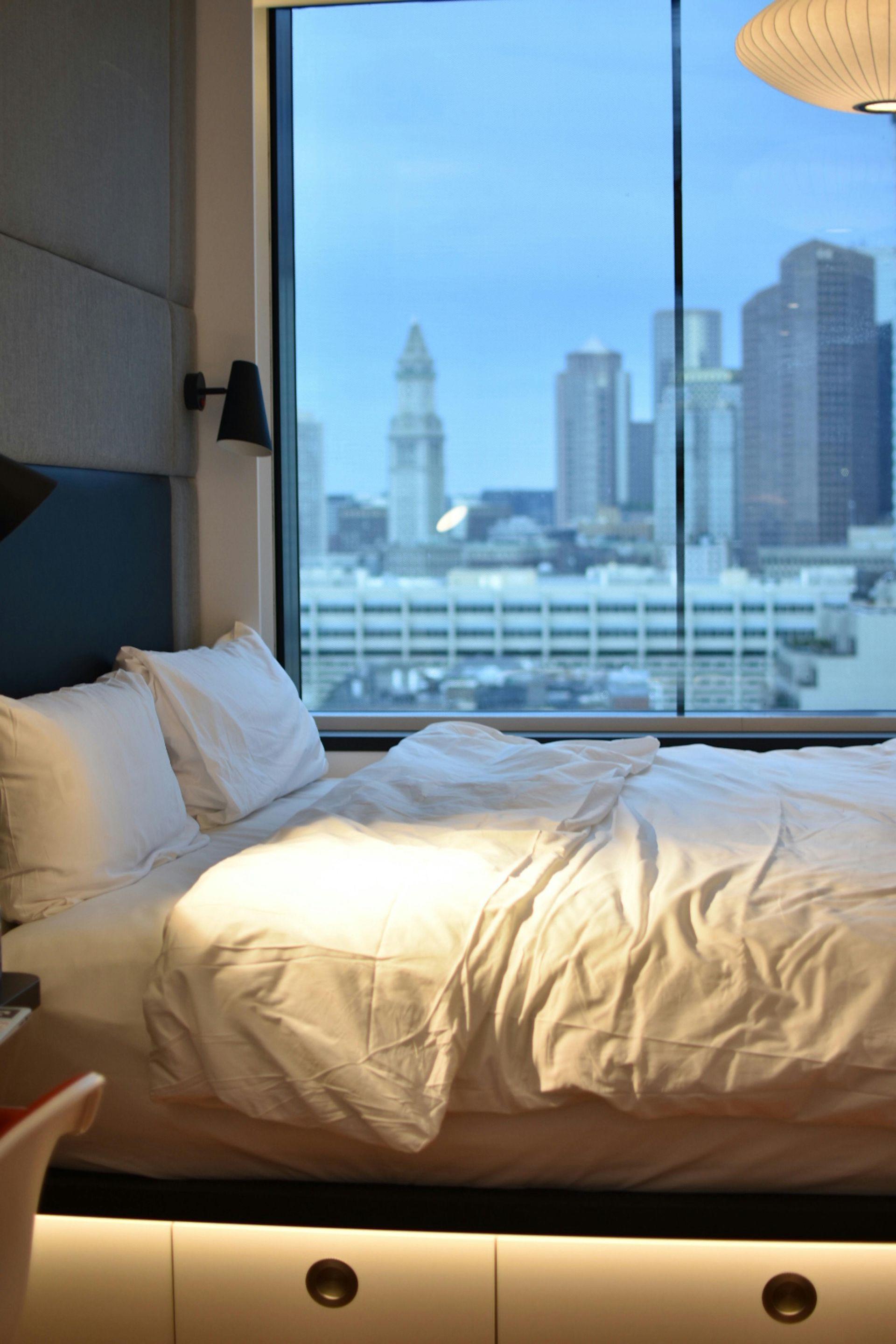 Cozy bed by a large window overlooking a city skyline at dusk