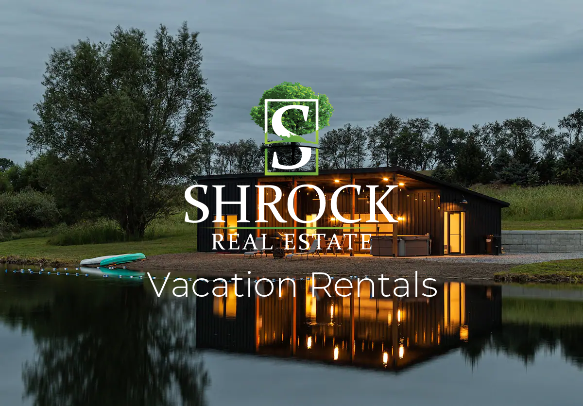 A beautiful house with lights on behind a lake. Shrock Real Estate Vacation Rentals logo in the foreground. 