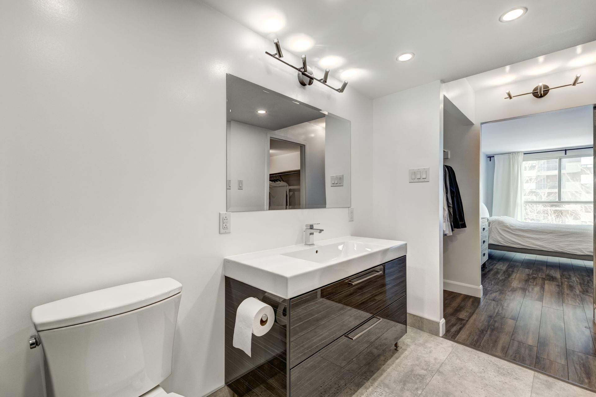 Modern bathroom with white walls, vanity, toilet, and doorway to a bedroom.