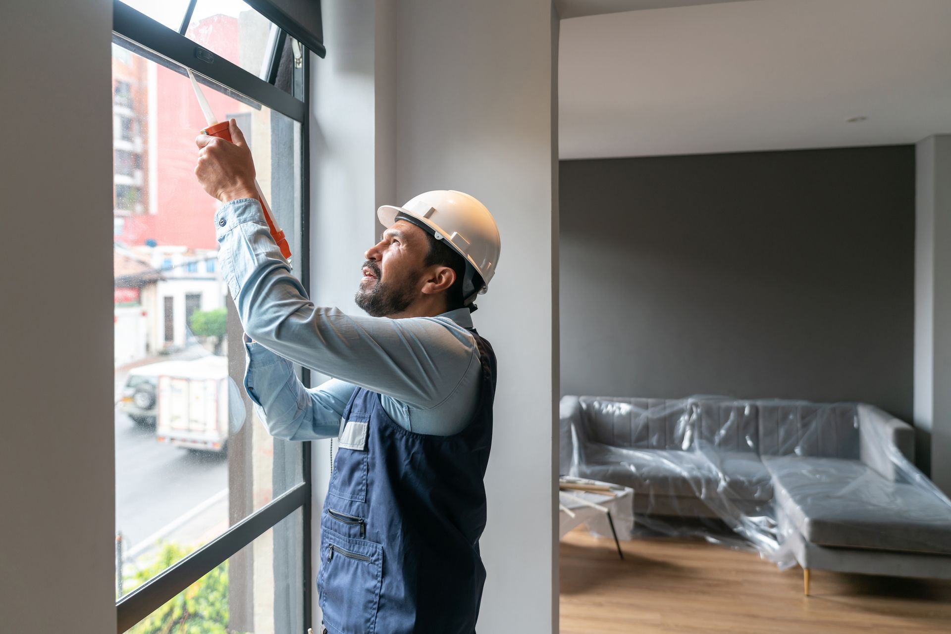 Construction worker, wearing a hard hat, caulking a window indoors.