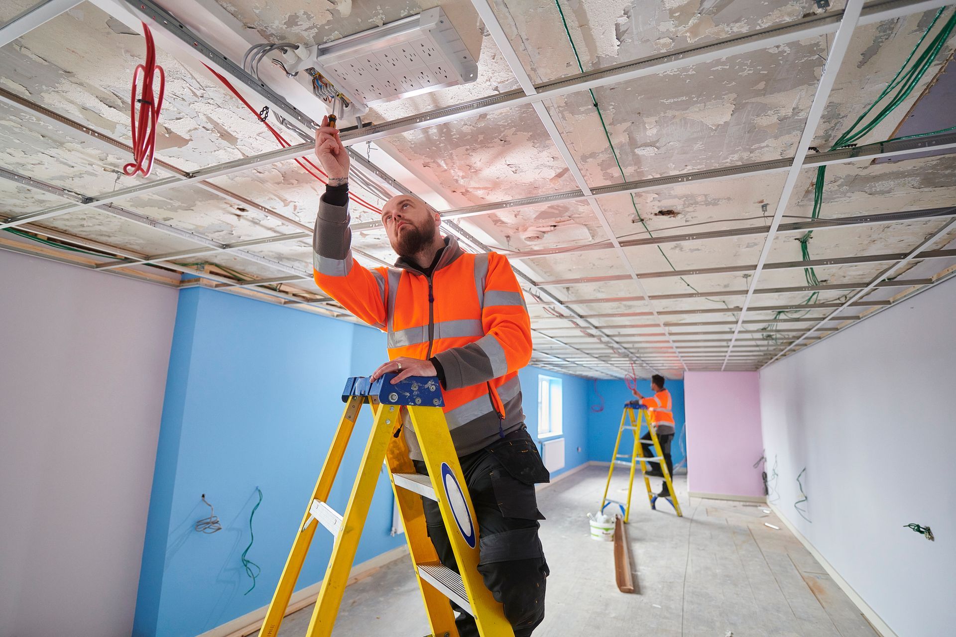 Construction worker on a ladder, installing ceiling tiles. Interior of a room under renovation, featuring exposed wiring.