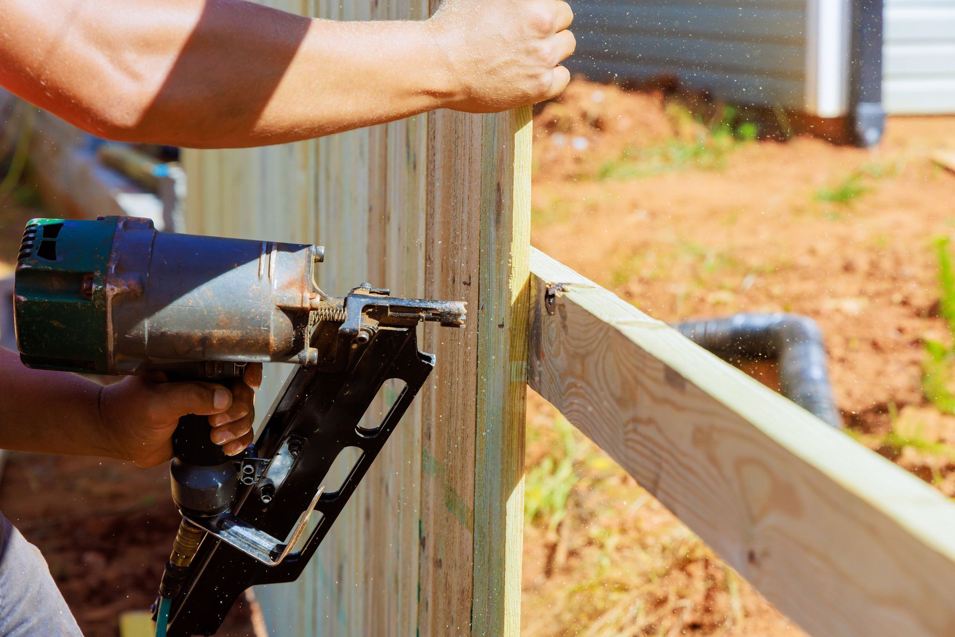 Person using a nail gun to attach a wooden fence board to a post outdoors.