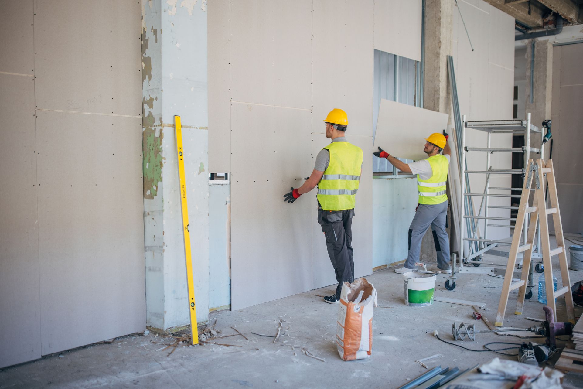 Construction workers installing drywall in a building. They are wearing safety vests and helmets.