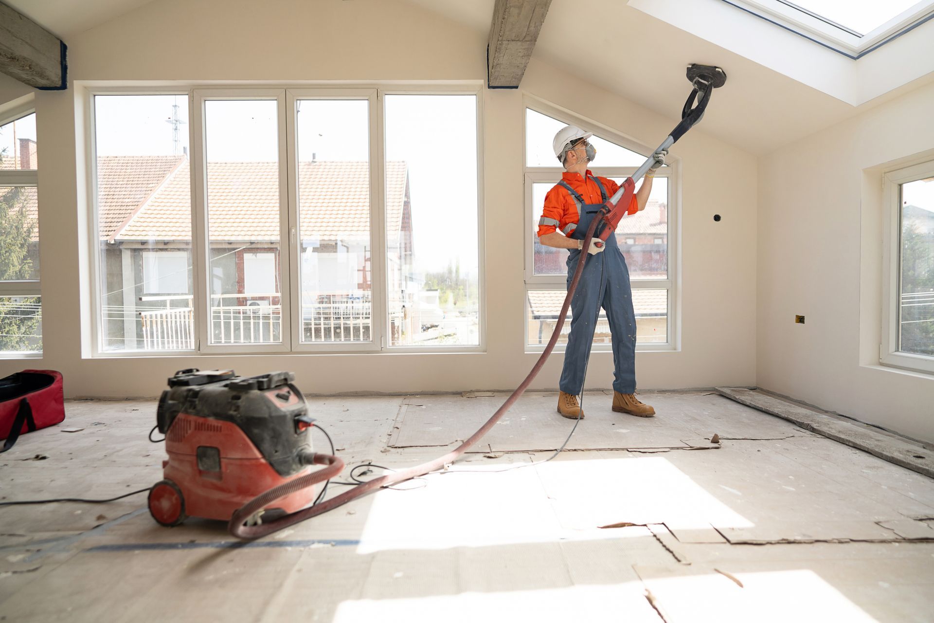 Person sanding ceiling with a sander, connected to a vacuum in a room with large windows.
