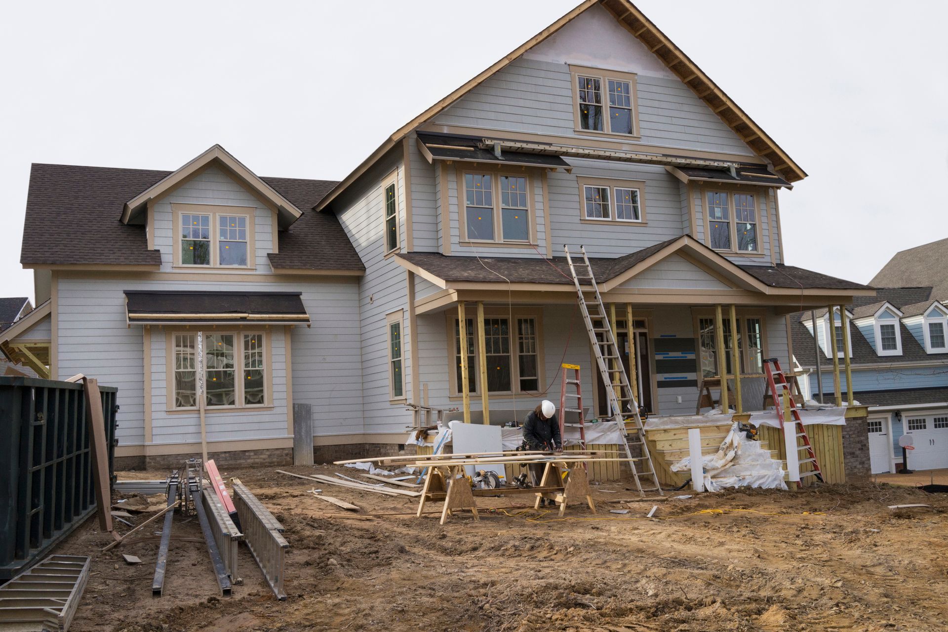 House under construction with worker on porch. Brown roof, light blue siding, ladders, and construction materials.