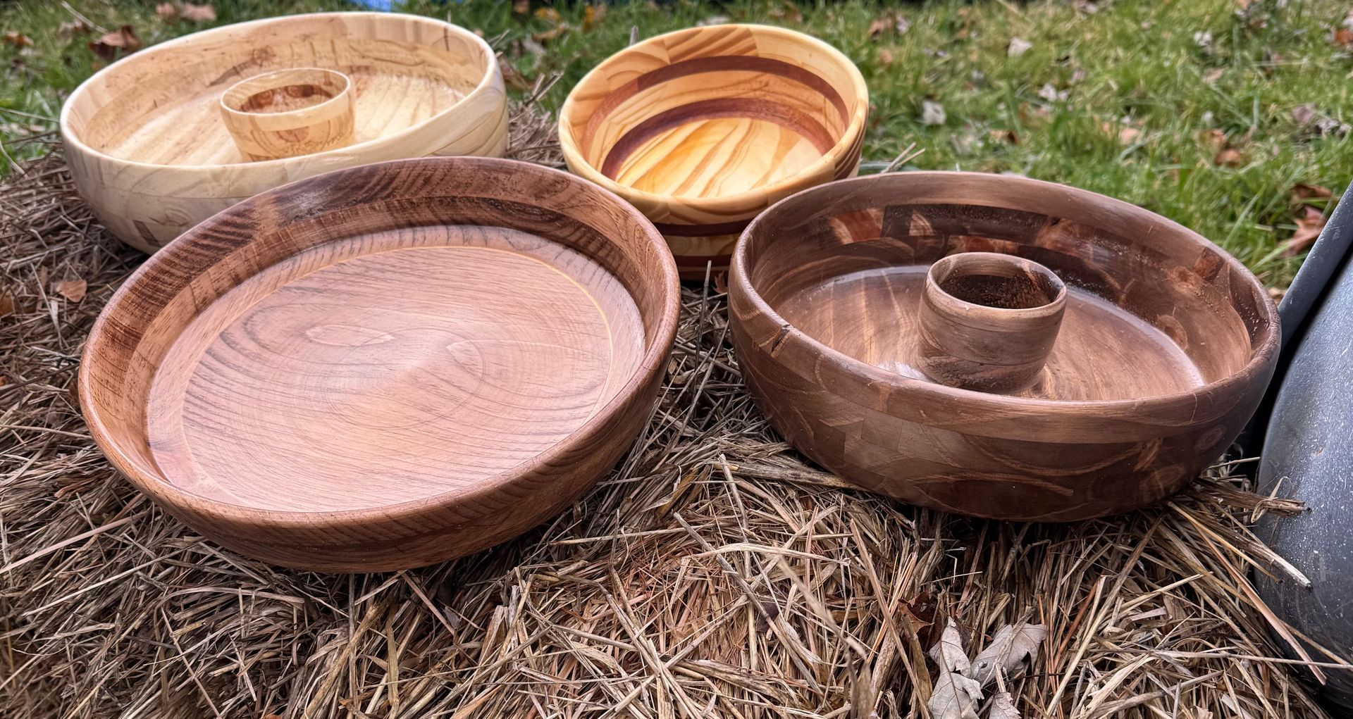 Four wooden bowls of varying shades, each with a central cup, rest on dry grass.