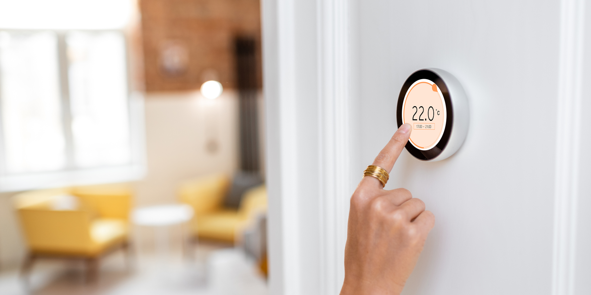 Hand pressing a doorbell on a white wall near a blurred, cozy living room with yellow chairs